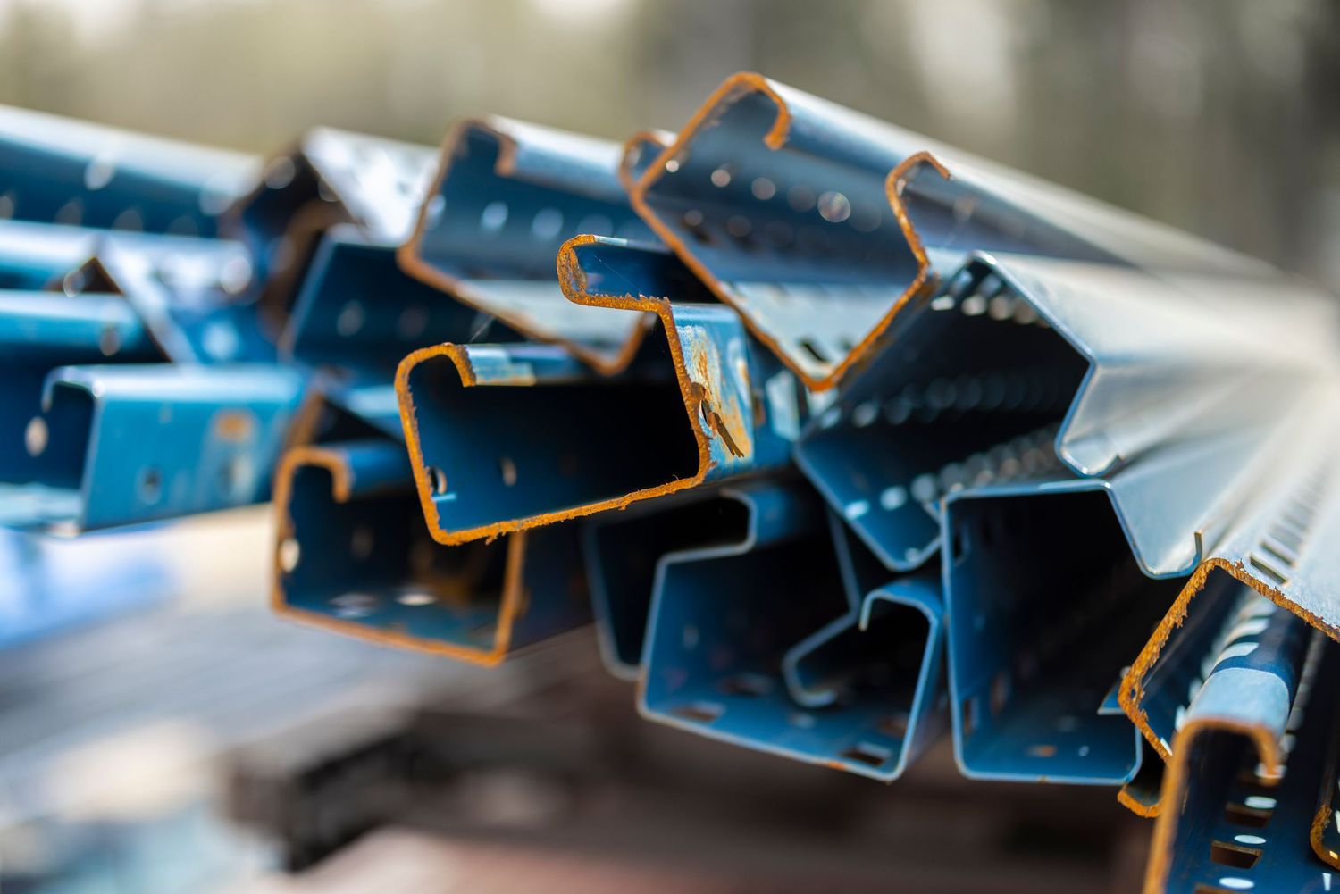 Stack of blue steel C-channel construction beams with rust spots stored outdoors at a building site.