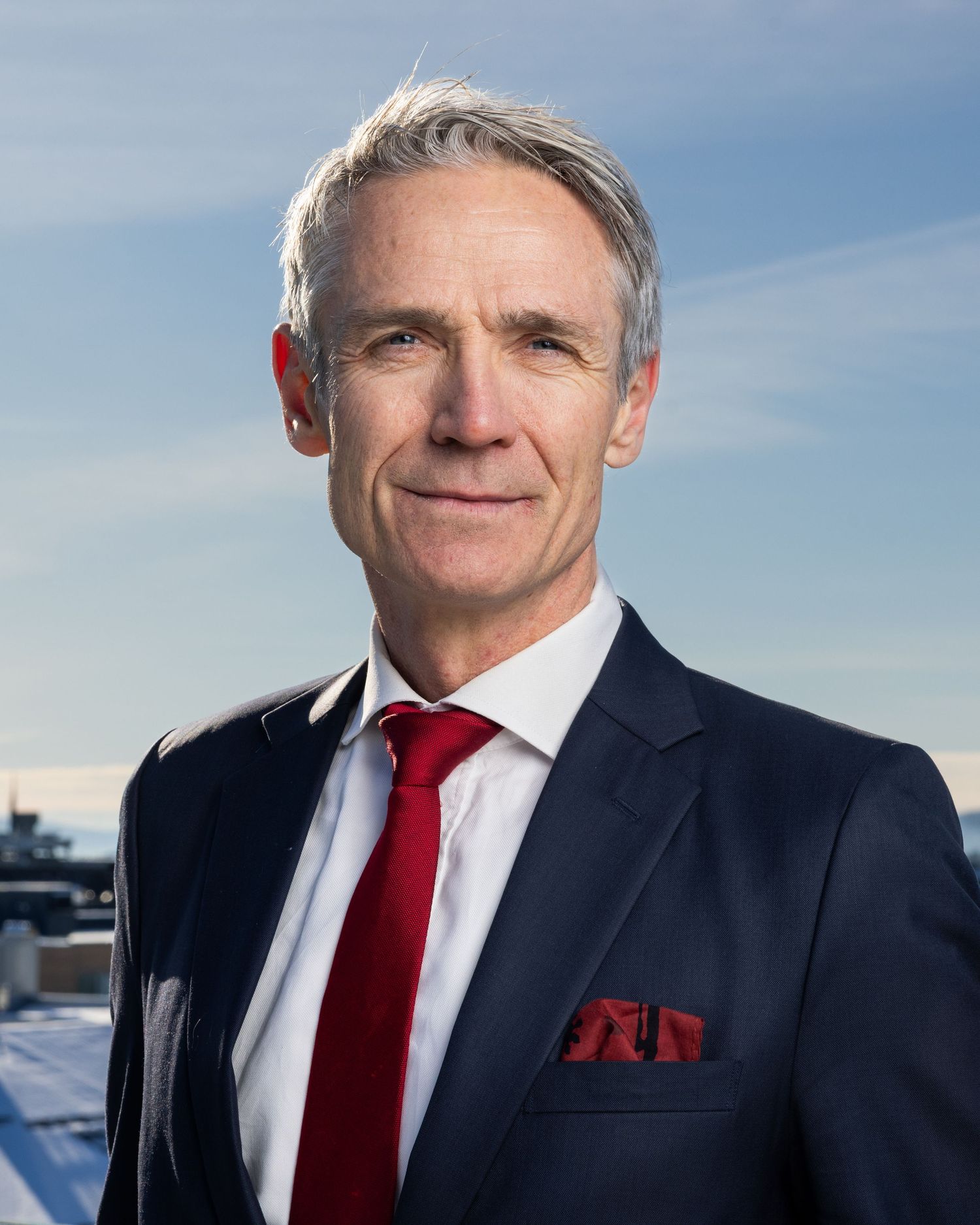 Professional older man in navy suit with red tie posing outdoors against a winter sky.