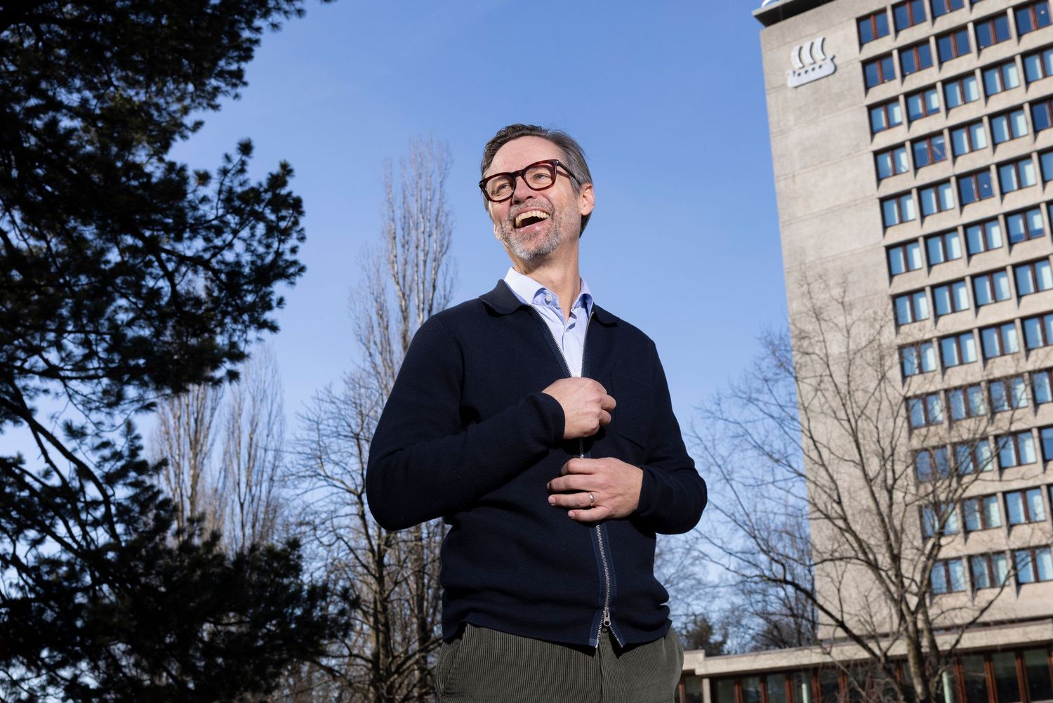 Smiling man in navy cardigan standing outdoors near a modern building on a sunny day.