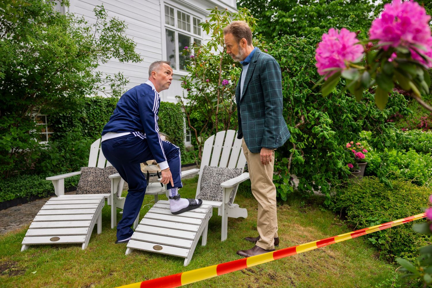 Two men examine white Adirondack chairs in a garden behind yellow crime scene tape.