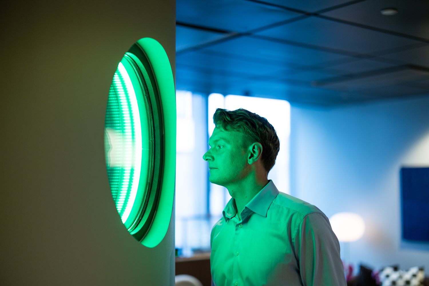 Man in a light green button-up shirt stands in profile gazing at a glowing circular green light panel in a modern office.