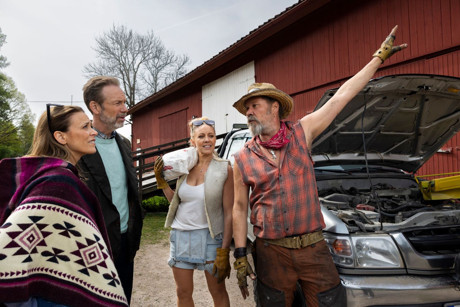 Four people laugh together near a red barn with an open car hood.