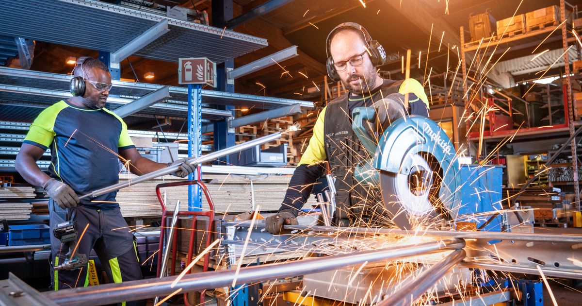 Two industrial workers grinding metal with angle grinder, sparks flying in busy manufacturing warehouse.