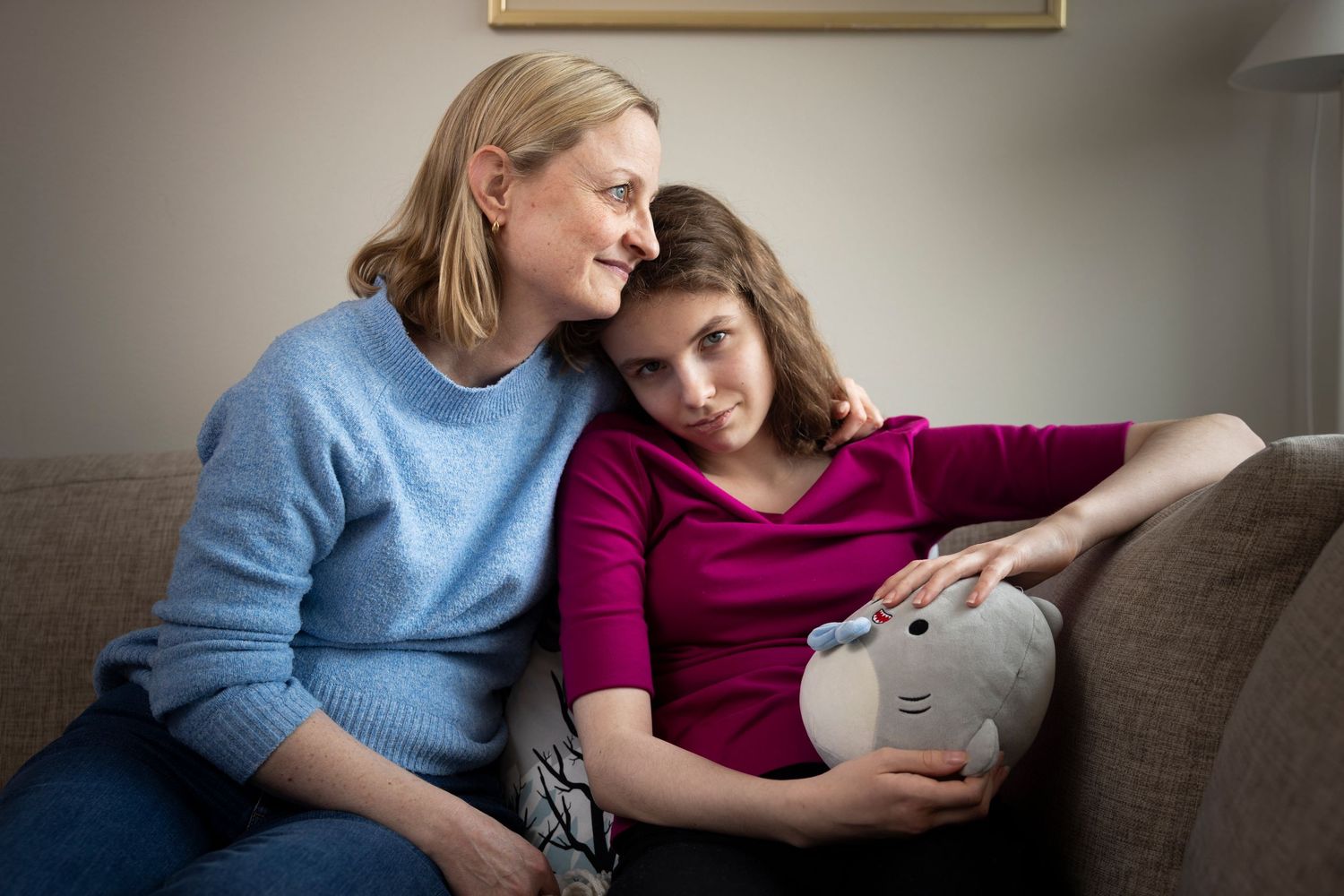 A mother comforts her teenage daughter on a couch, the girl holding a stuffed animal.