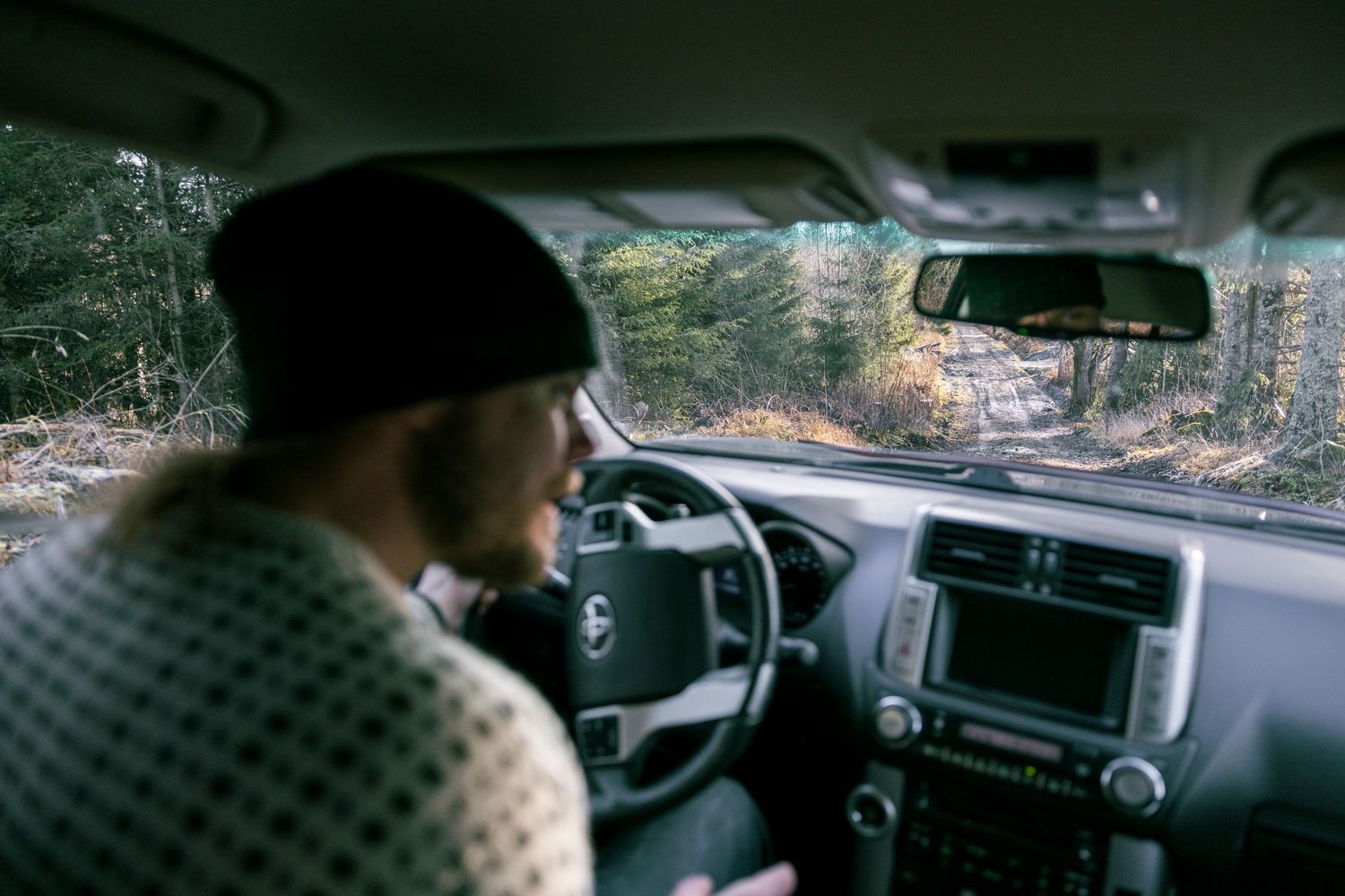 Man in beanie hat driving an SUV through a forest road viewed from back seat.