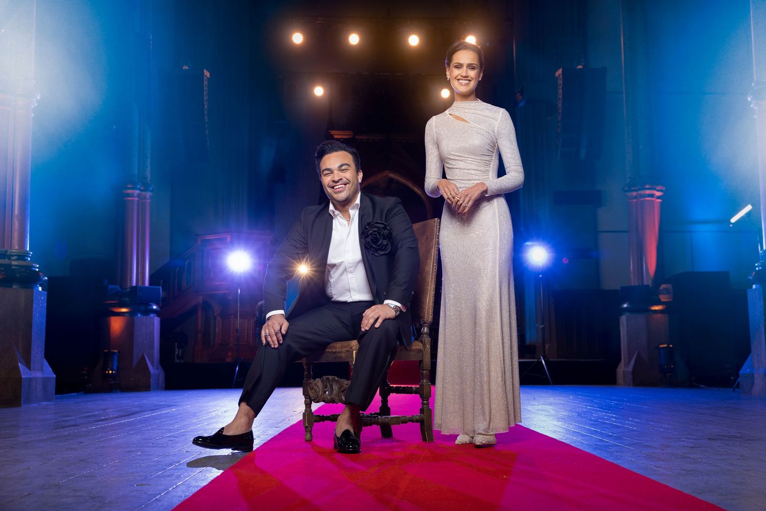 Elegant man and woman in formal attire pose on a red carpet in a dramatic lit venue.