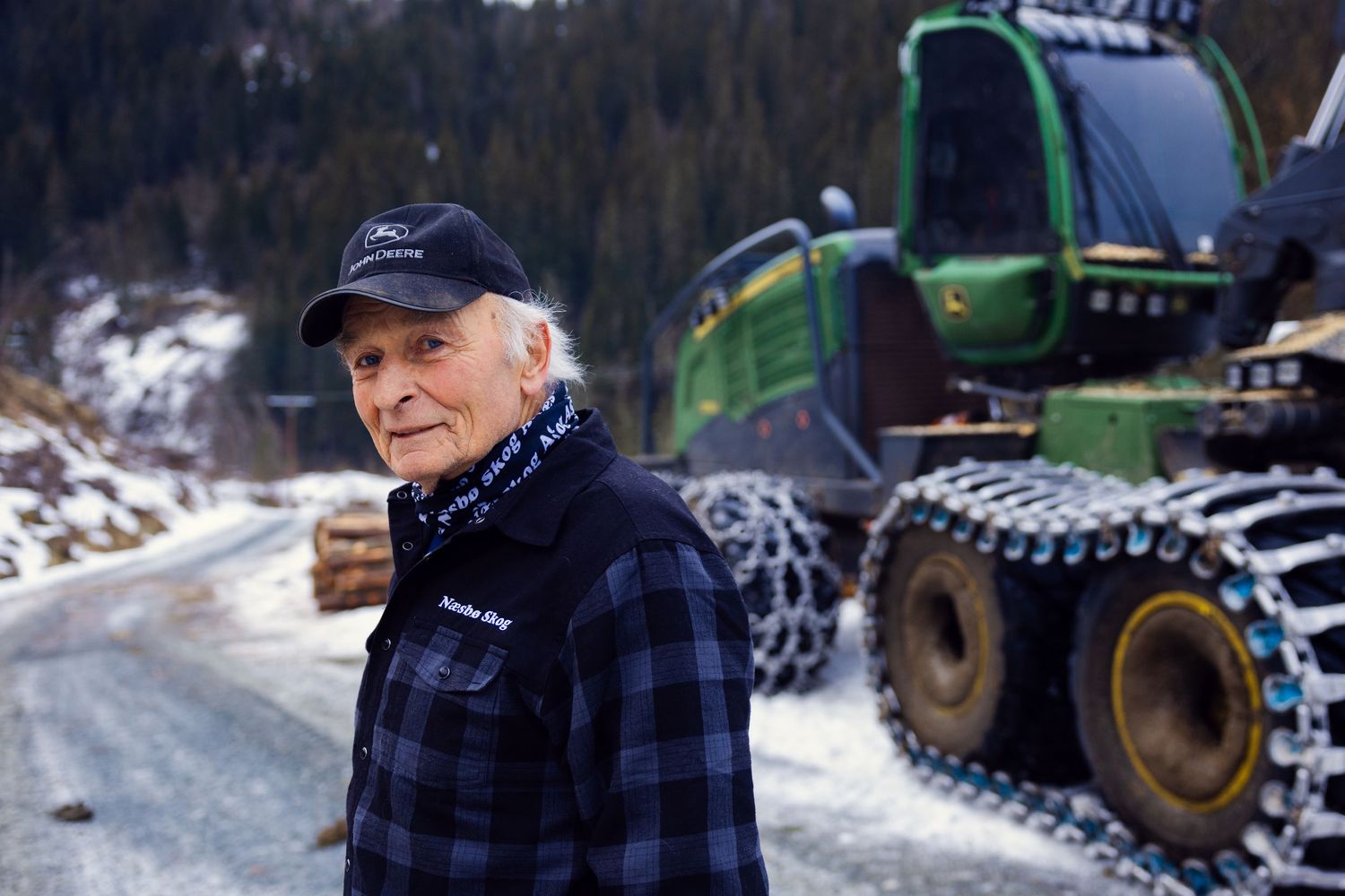 Elderly man in plaid shirt standing before a green John Deere forwarder on a snowy road.