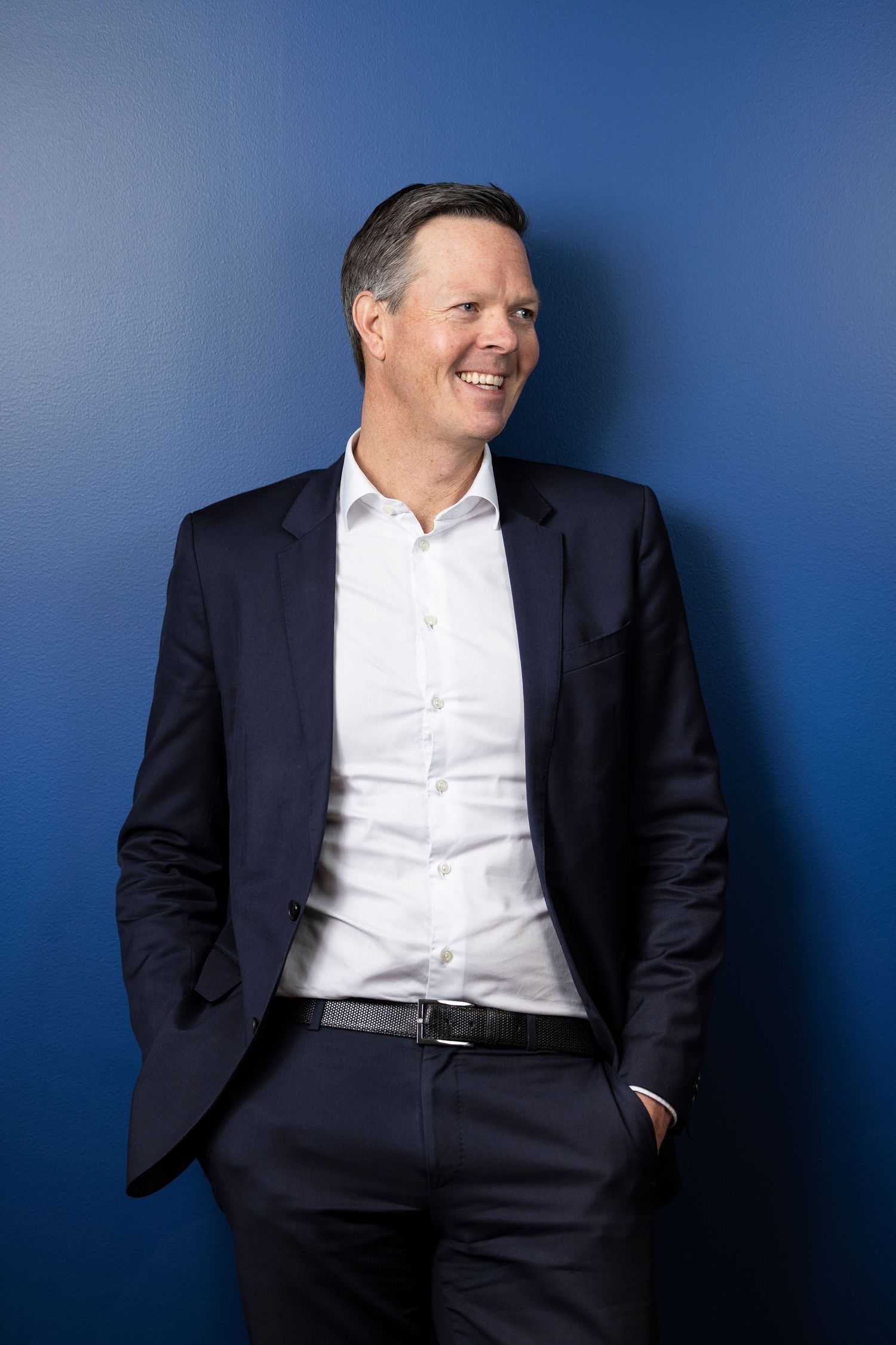 A smiling businessman in a dark suit and white shirt poses confidently against a blue background.