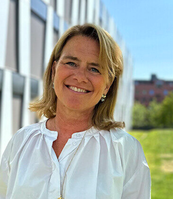 Smiling blonde woman in a white blouse standing outdoors near a modern building on a sunny day.