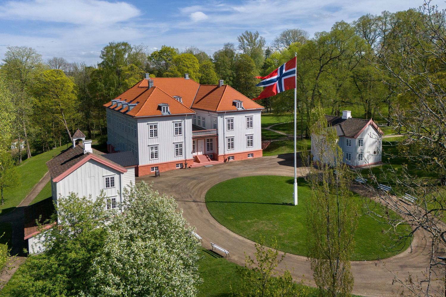 Aerial view of a historic Norwegian manor house with red roof and Norwegian flag in lush green surroundings.
