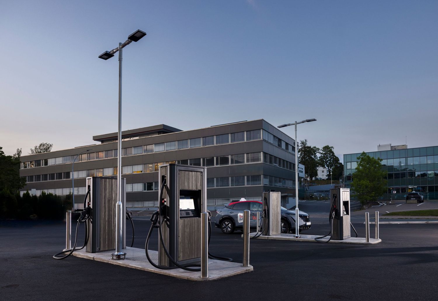 Modern electric vehicle charging station in empty parking lot at dusk with office building backdrop.