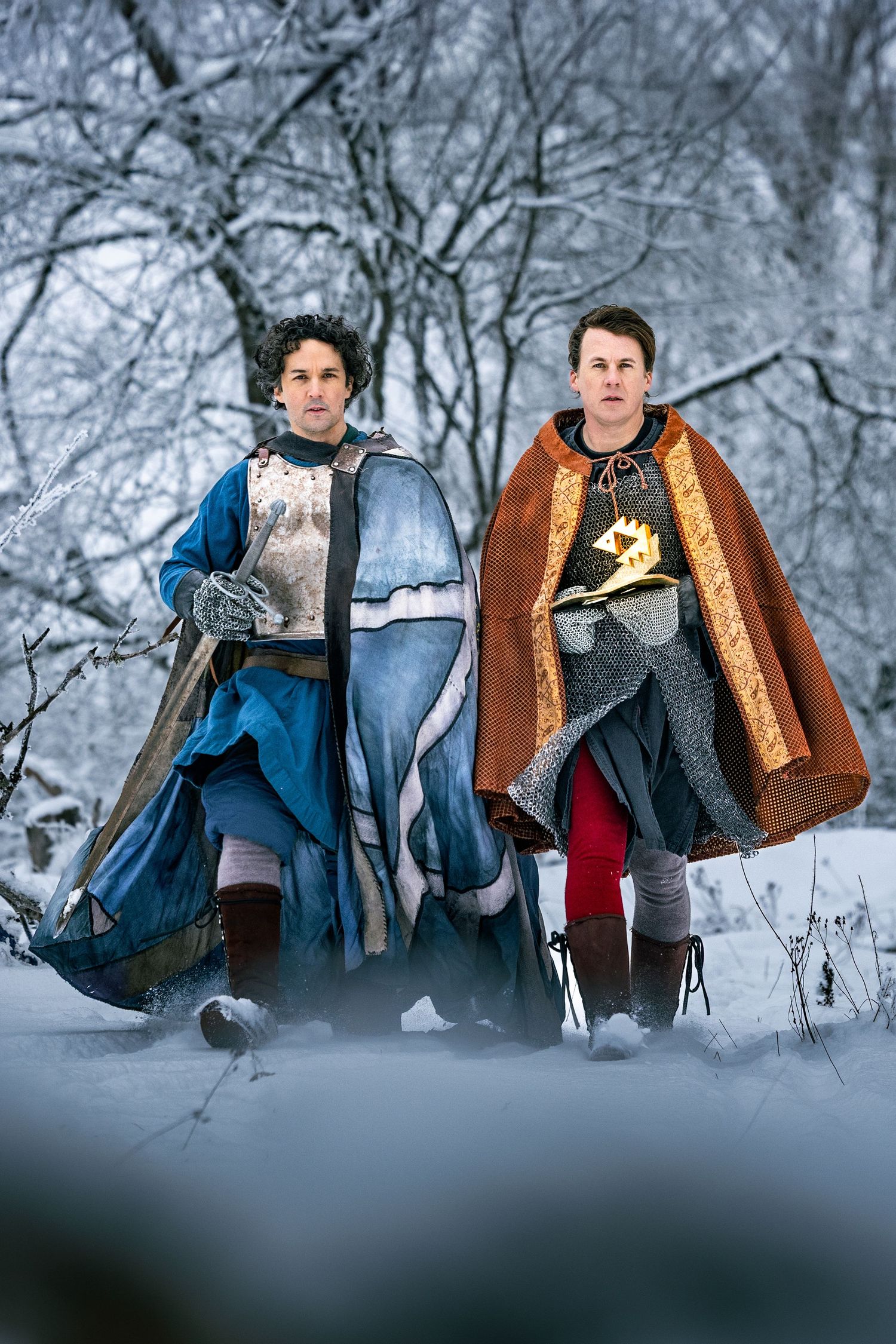 Two men dressed in knight armor and colorful capes walking through a snowy forest.