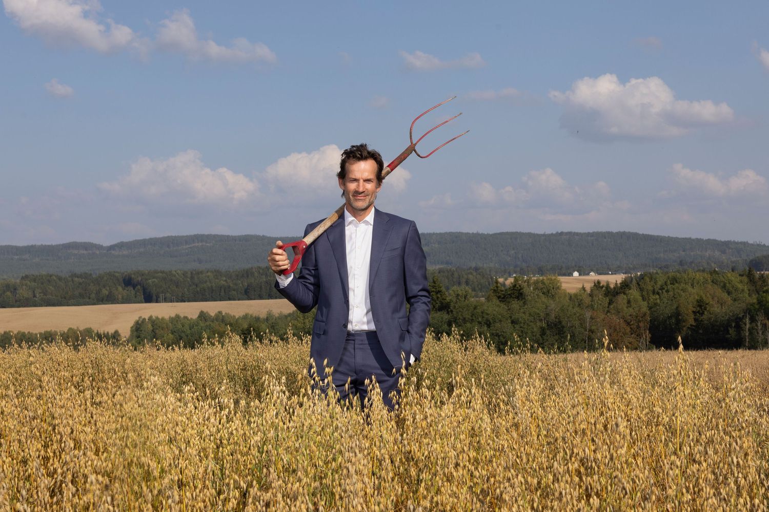 Smiling man in blue suit holding a red pitchfork standing in a golden grain field.