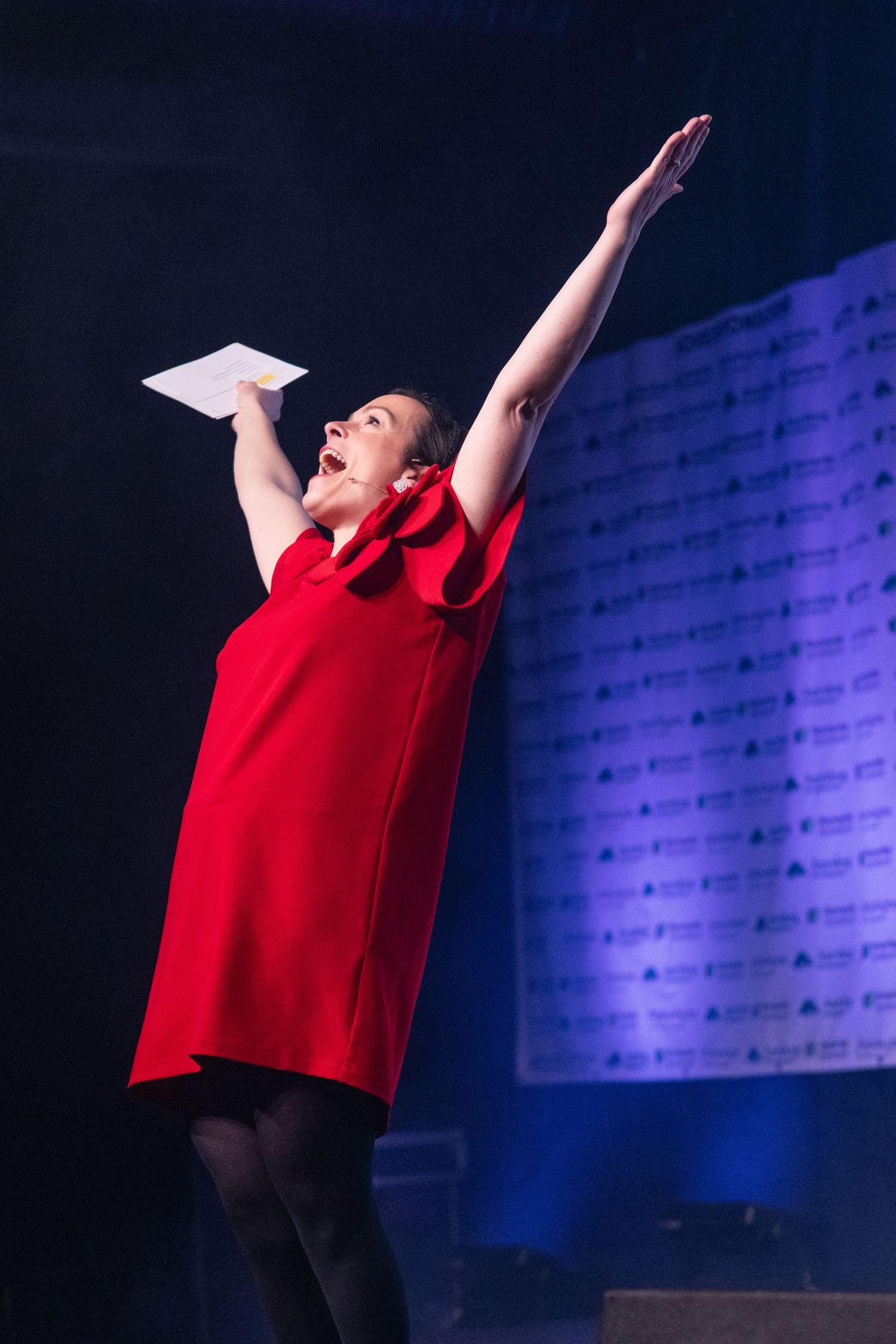 Woman in red dress raises arm dramatically on stage while holding papers at a live event.