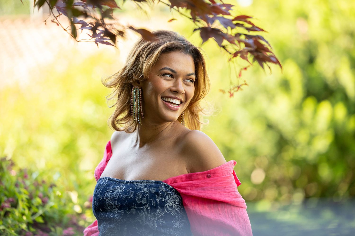 Smiling woman in pink wrap dress poses under colorful foliage in sunlight.