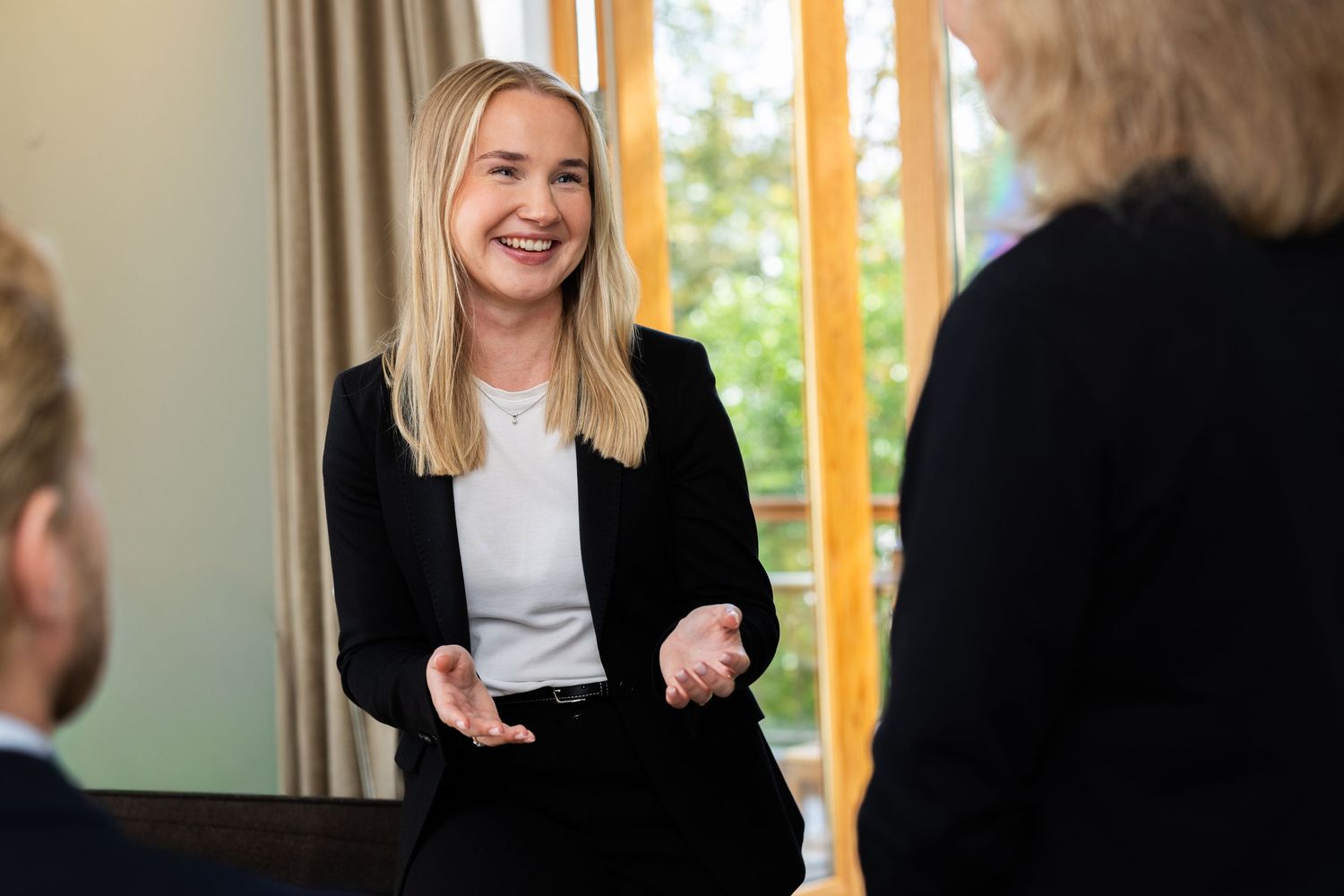 Young blonde woman in black blazer smiling and gesturing during professional business meeting.