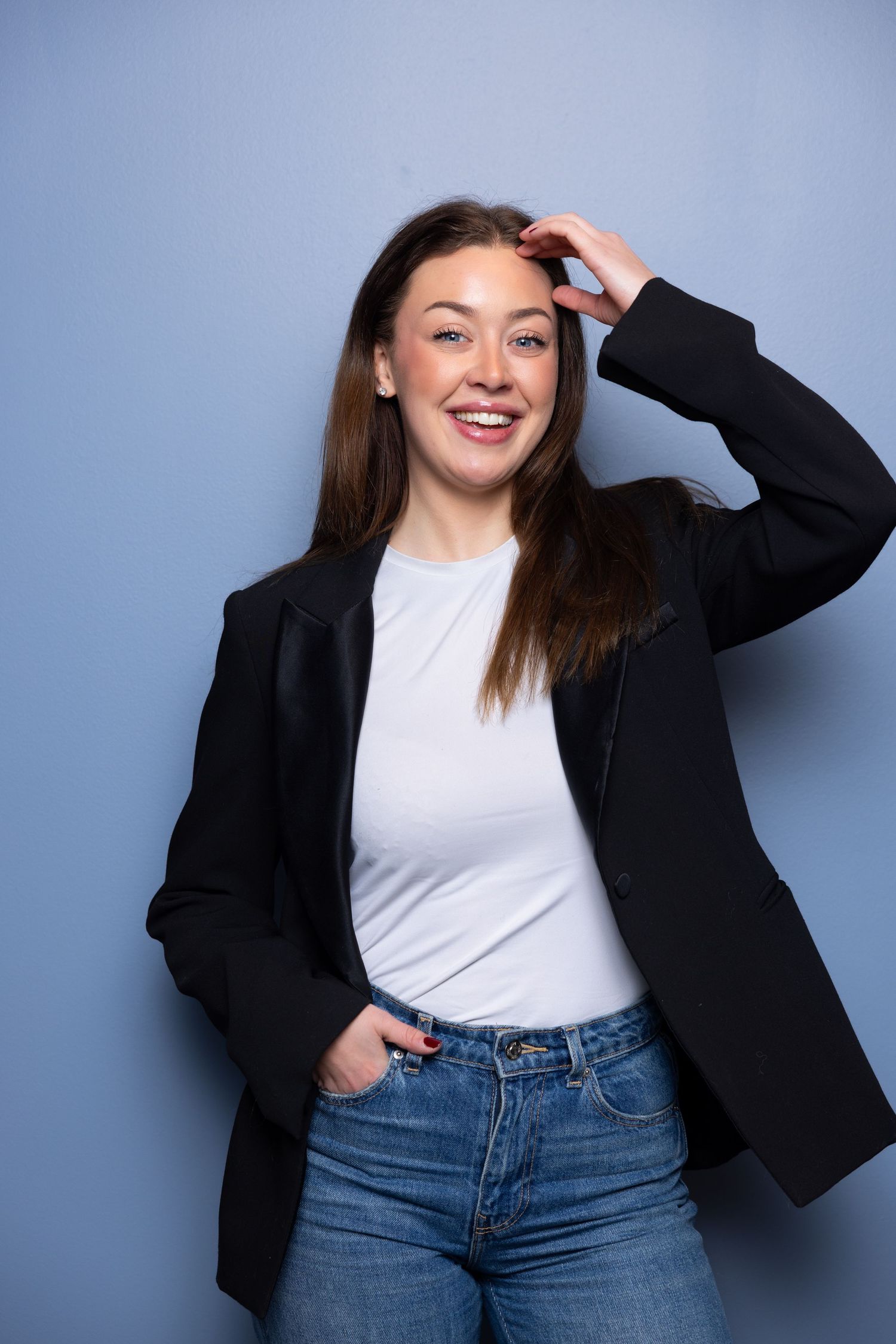 Smiling young woman in black blazer, white tee, and jeans posing against a blue background.