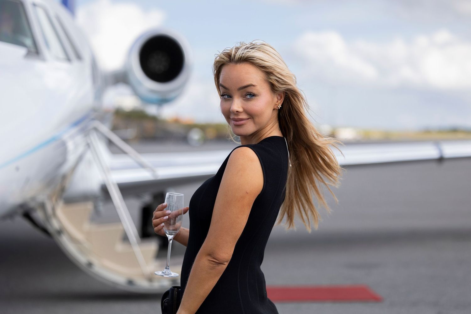 Stylish blonde woman in black dress holding champagne glass outside a private jet on tarmac.