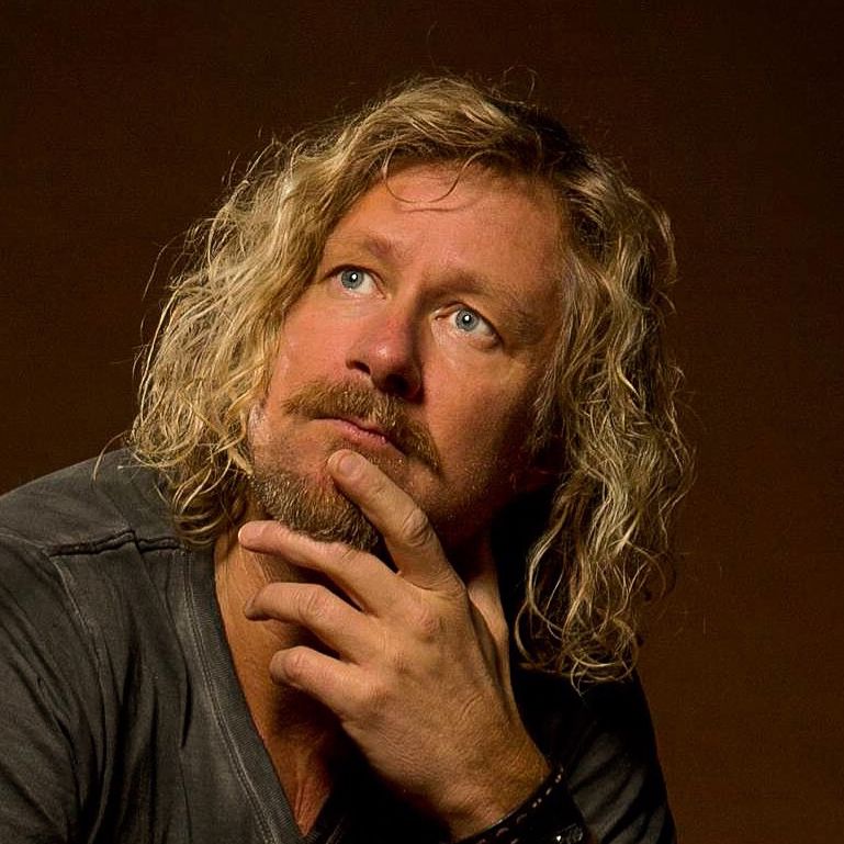 Man with long blonde curly hair and beard posing thoughtfully against a dark background.