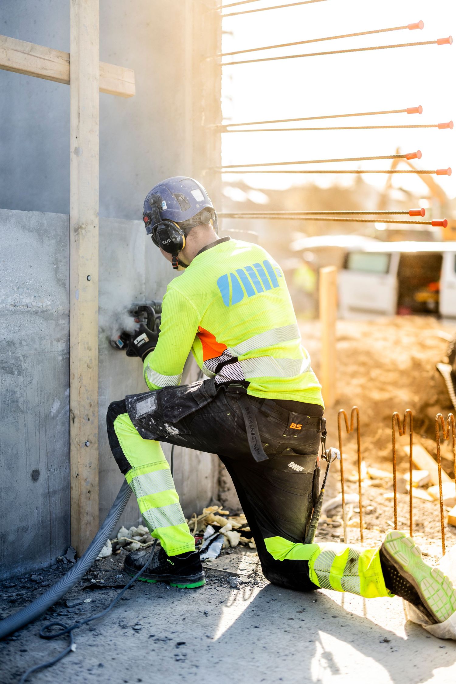 Construction worker in yellow hi-vis gear kneeling while drilling into a concrete wall at a building site.