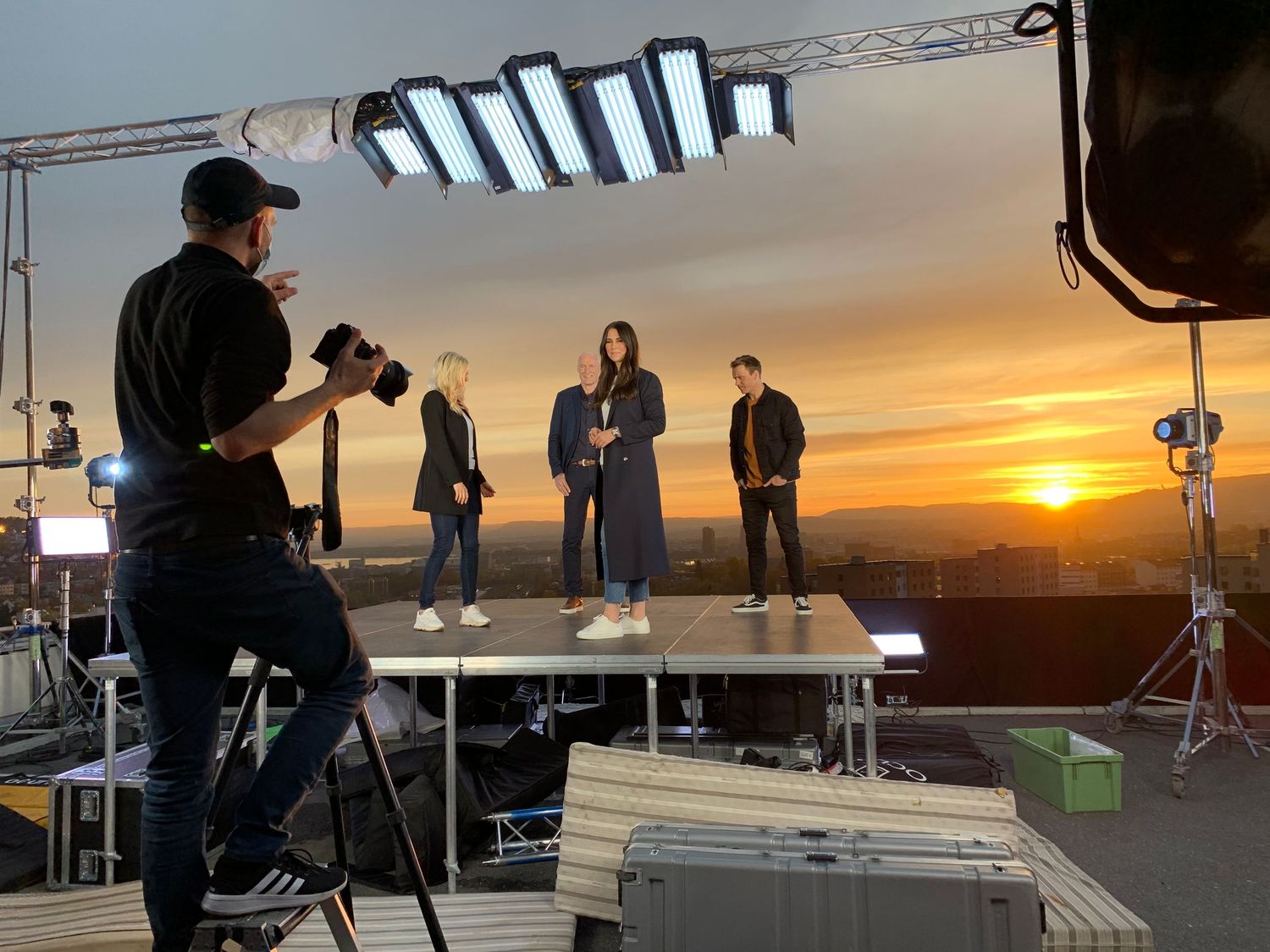 Photographer shoots three models on a rooftop stage with professional lighting equipment against a stunning sunset backdrop.