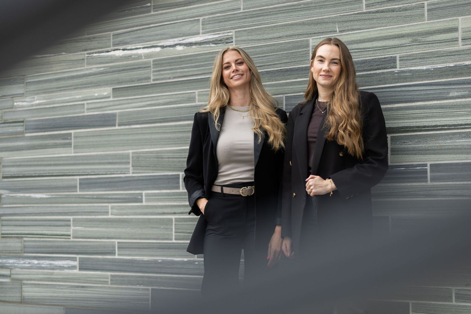 Two women with long hair in dark blazers stand side by side against a gray striped stone wall, smiling softly at the camera.