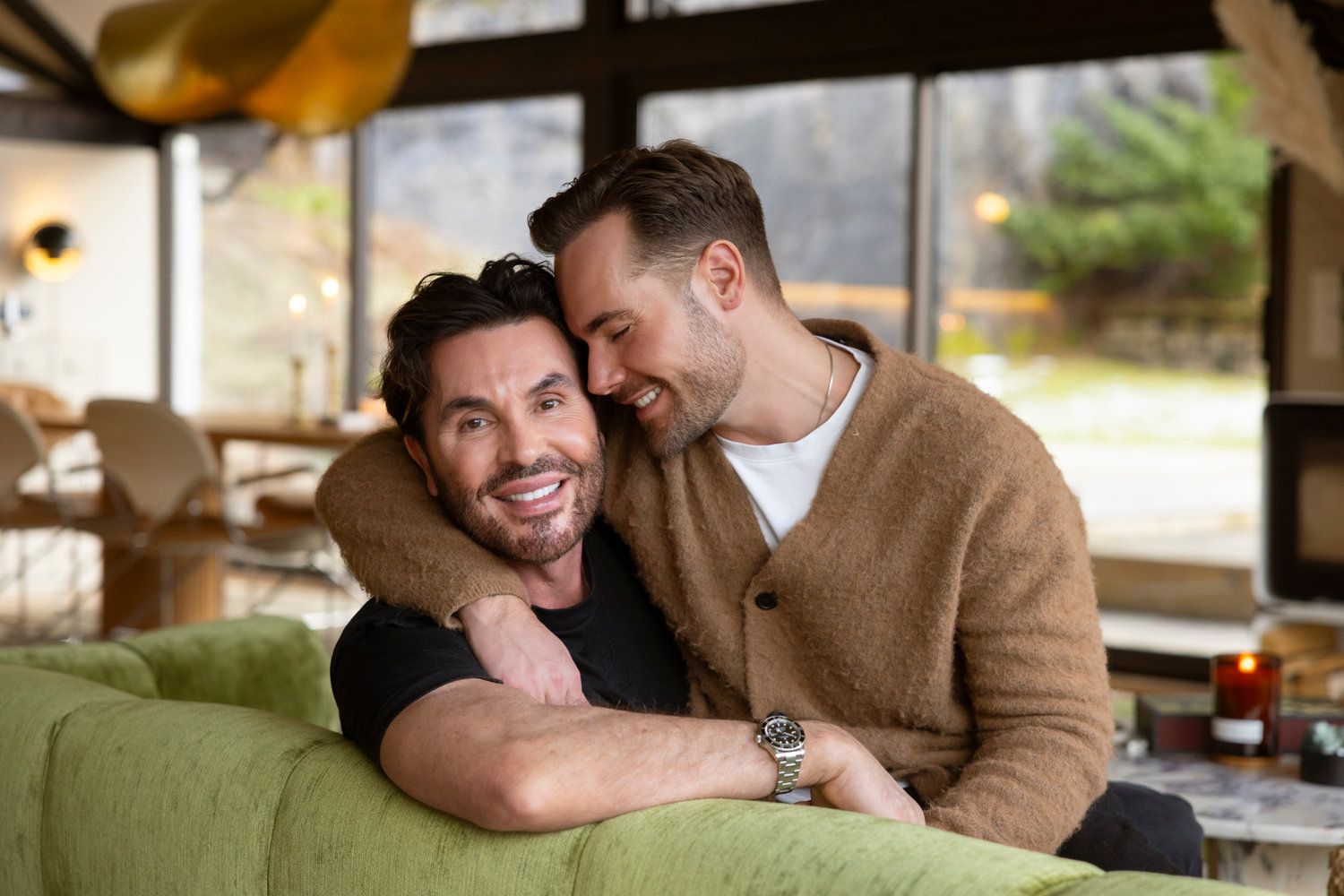 Two smiling men sharing a warm embrace on a green couch in a cozy living room.