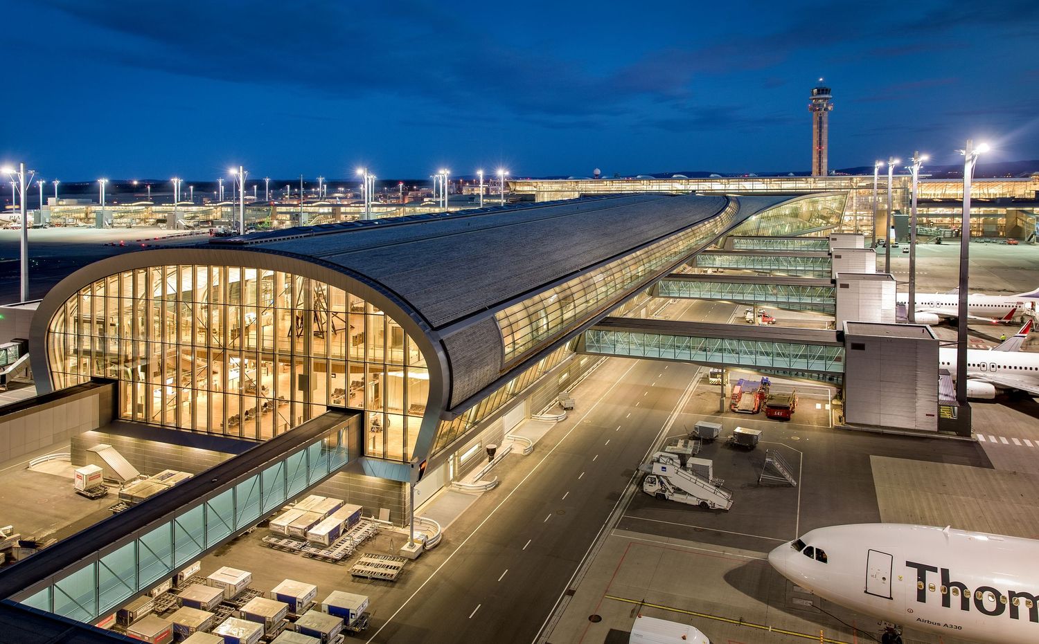Aerial night view of a large international airport terminal with planes docked and city lights in the background.