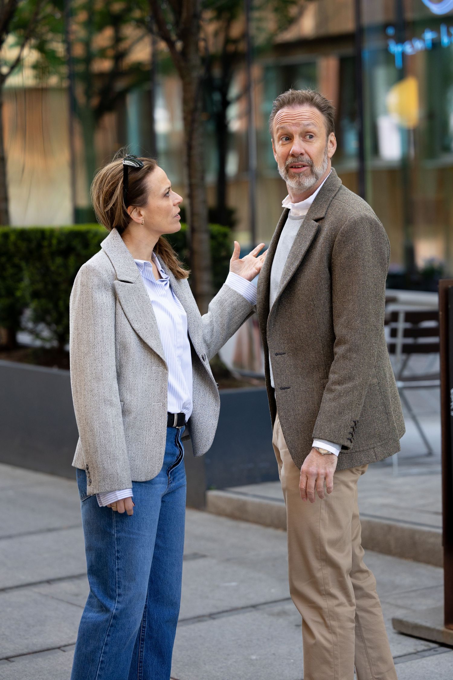 Woman in blazer gesturing while speaking to tall bearded man on urban city street.