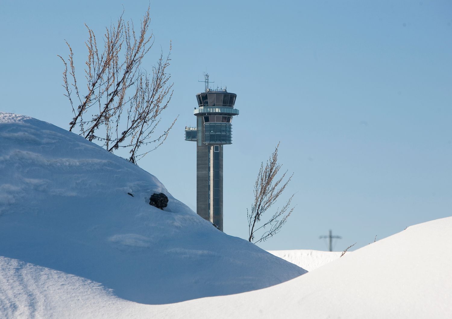 Snow-covered winter landscape with an airport control tower visible behind large white snowdrifts under clear blue sky.
