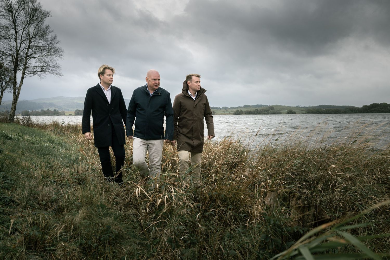 Three men walk together along a scenic lakeside with overcast skies and tall grass.
