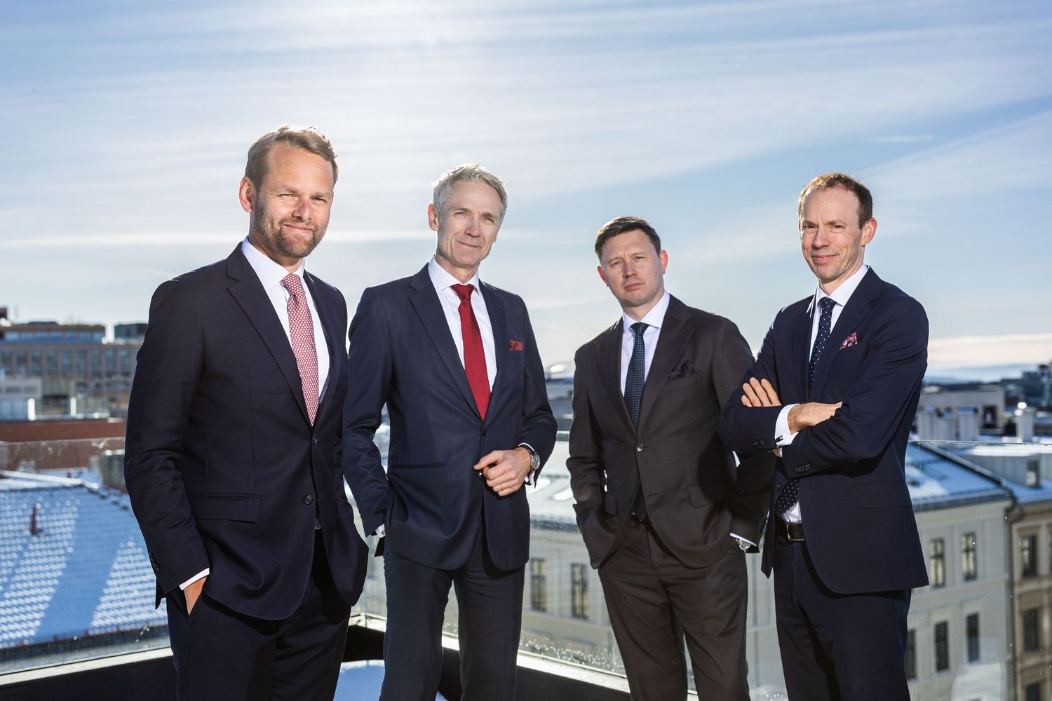 Four professional businessmen in suits posing together on a rooftop with city skyline behind them.