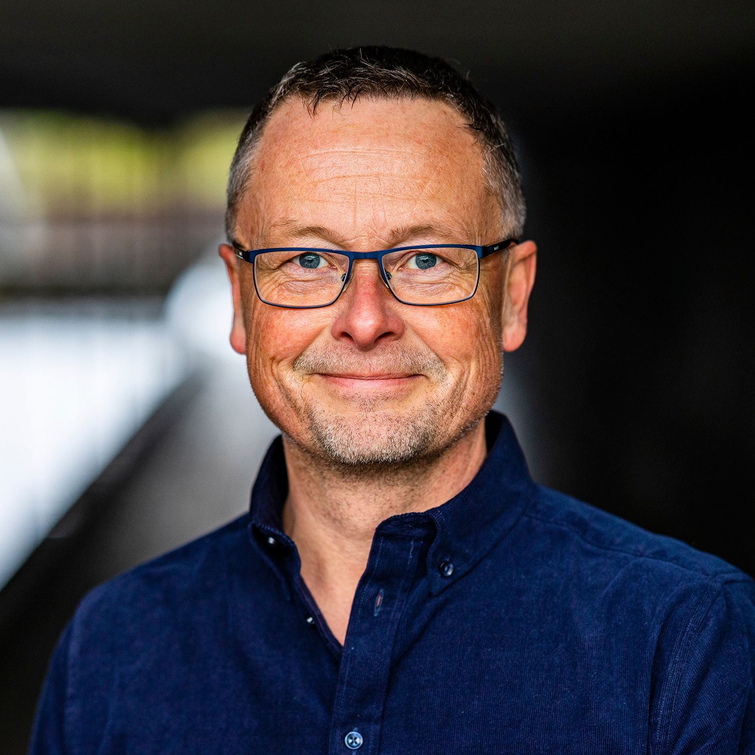Smiling middle-aged man with glasses wearing a dark blue shirt in a modern building.