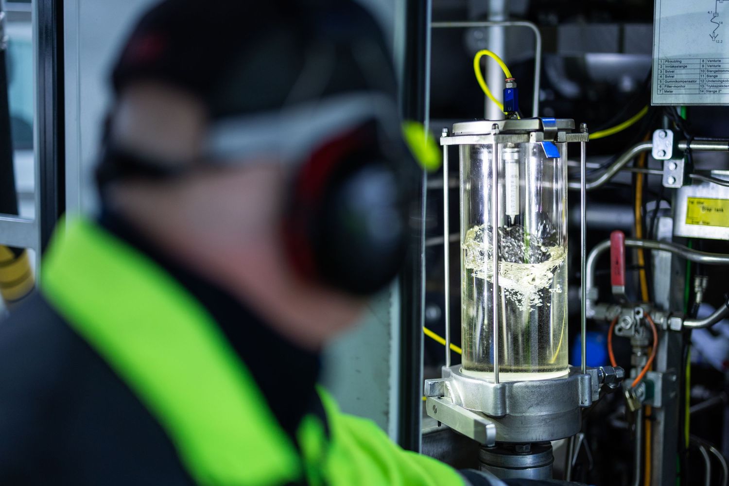 Technician in safety earmuffs inspects industrial glass cylinder equipment in a manufacturing facility.