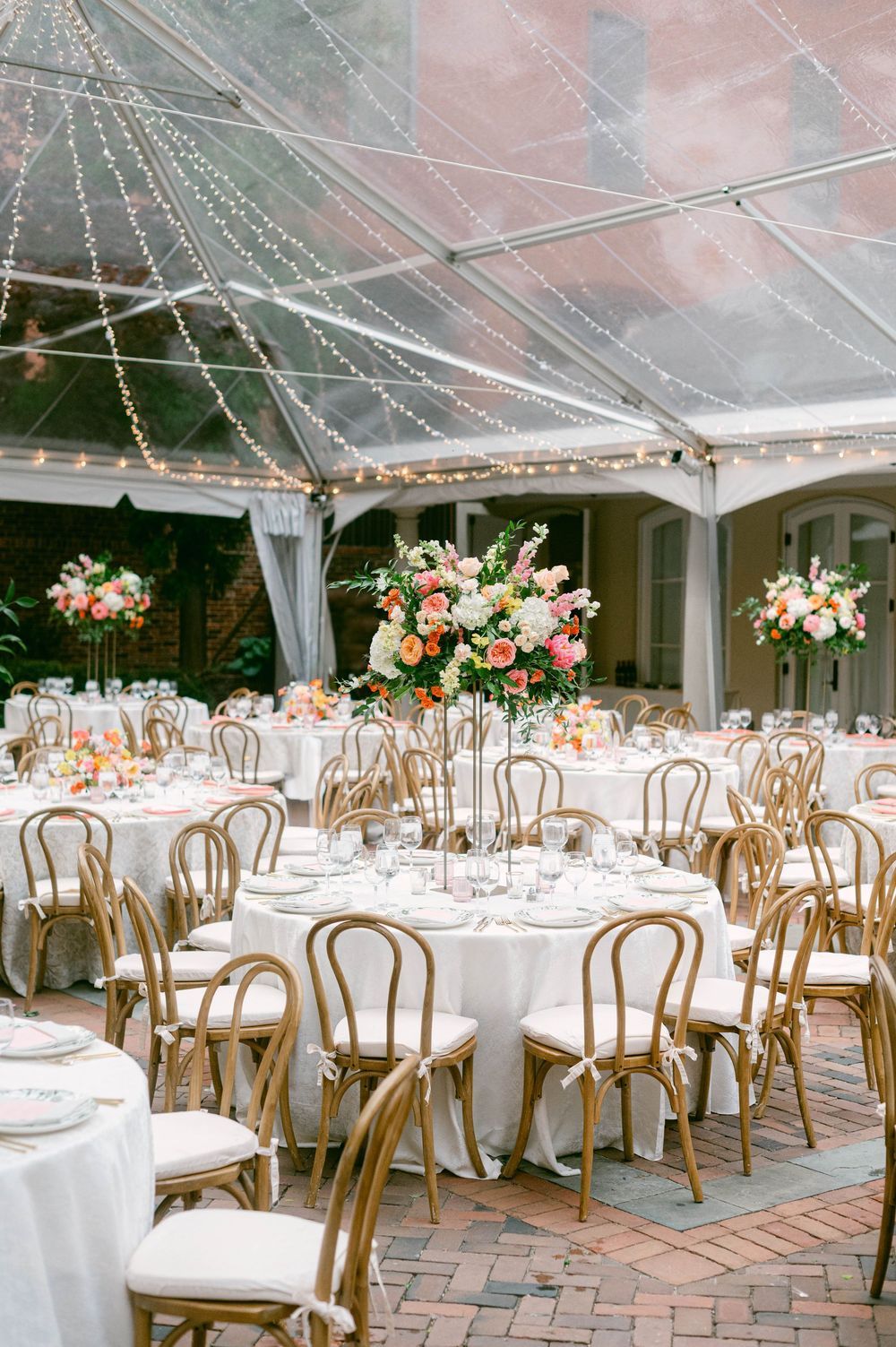 Elegant wedding reception setup with round tables, wooden chairs, and floral centerpieces under a clear tent.