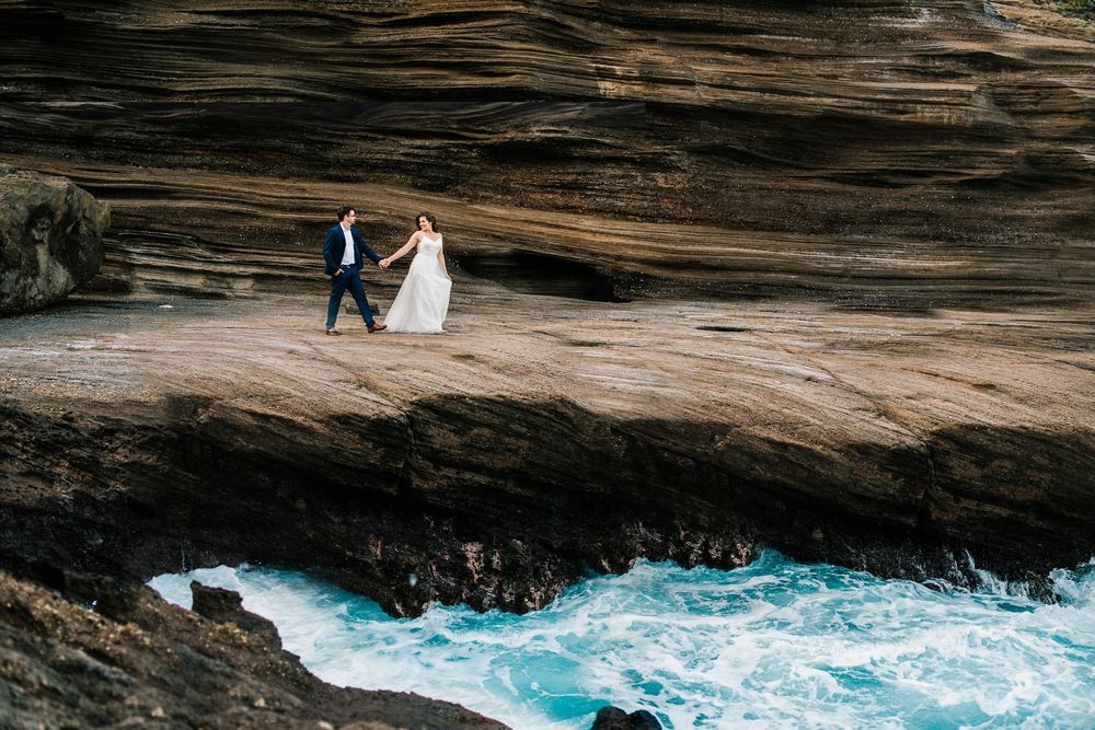 Couple stands near dramatic cliffside cave overlooking turquoise ocean waves crashing against rocky shoreline.
