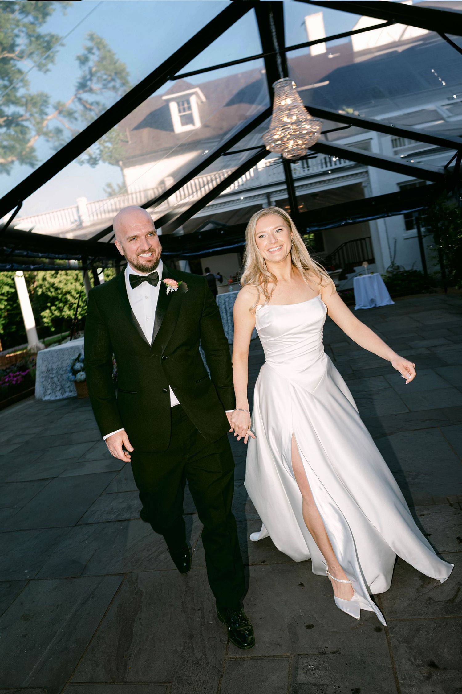 Smiling bride in white gown and groom in black tuxedo hold hands walking under glass canopy at elegant outdoor wedding venue.