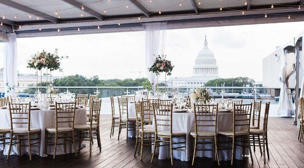 Panoramic view of an elegant wedding reception venue with white chairs and tables set up beneath a tented ceiling.