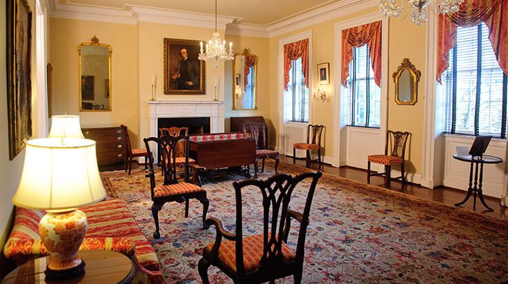Panoramic view of an elegant Victorian-style parlor room with antique furniture, red carpeting and ornate window treatments.