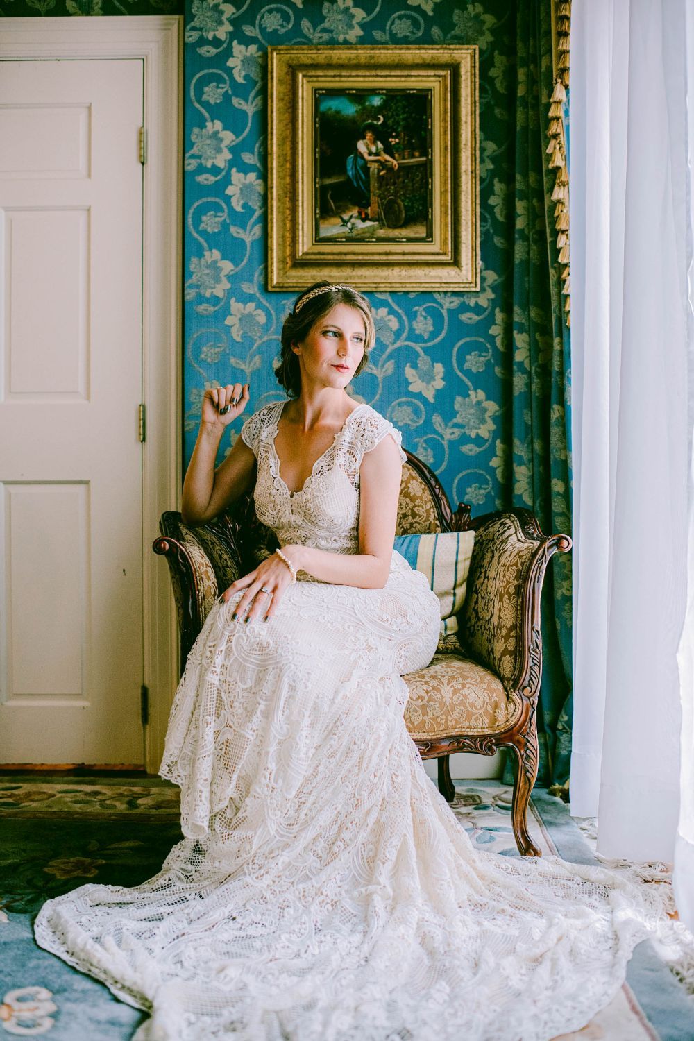 Elegant bride in lace wedding gown poses on vintage chair against blue damask wallpaper in classic interior setting.
