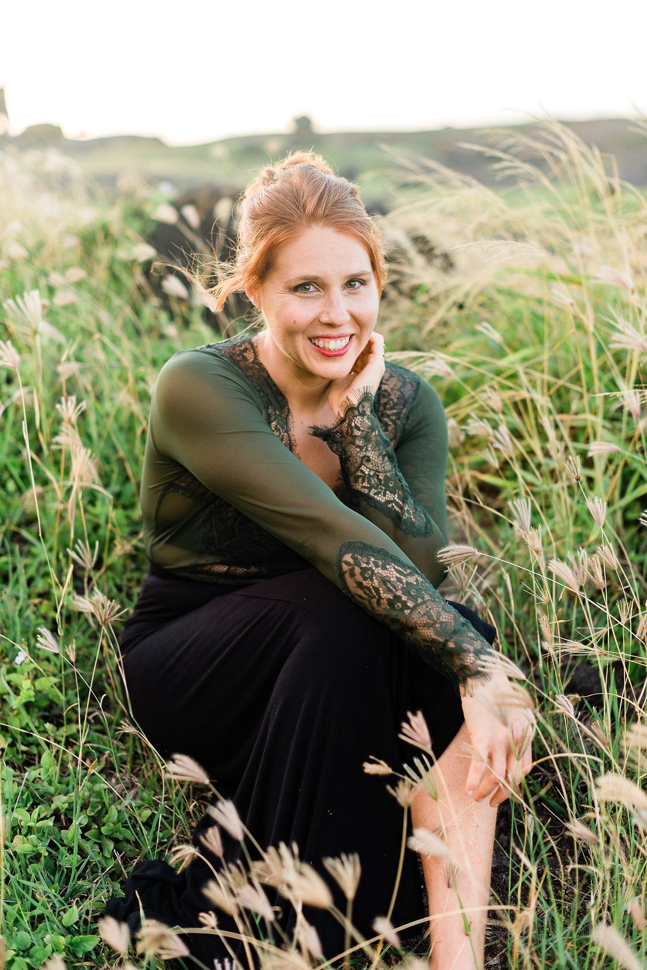 A person in a dark dress and green top poses gracefully in a sunlit meadow of tall grass and wildflowers.