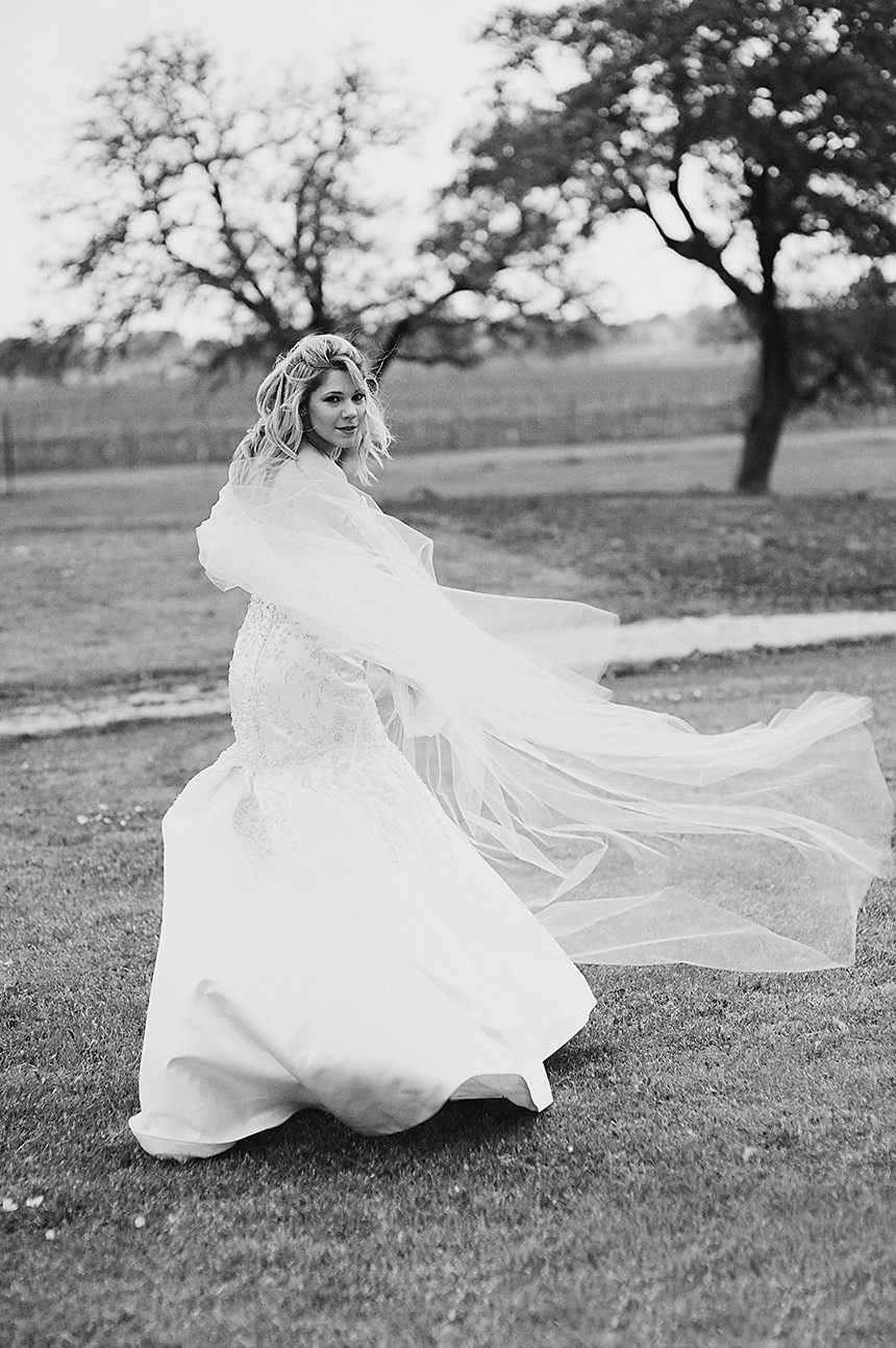 A bride in a flowing white wedding dress twirls on a grassy field with trees in the background, captured in black and white.