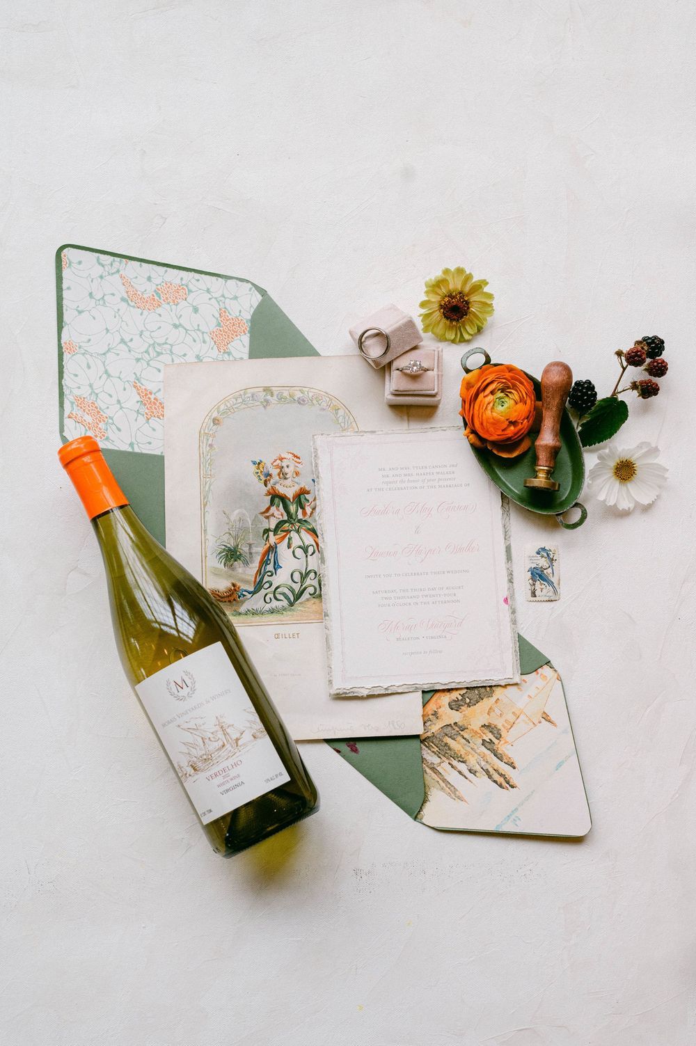 Flatlay of wedding stationery with vintage illustration, wine bottle, small pumpkin and floral accents on white surface.