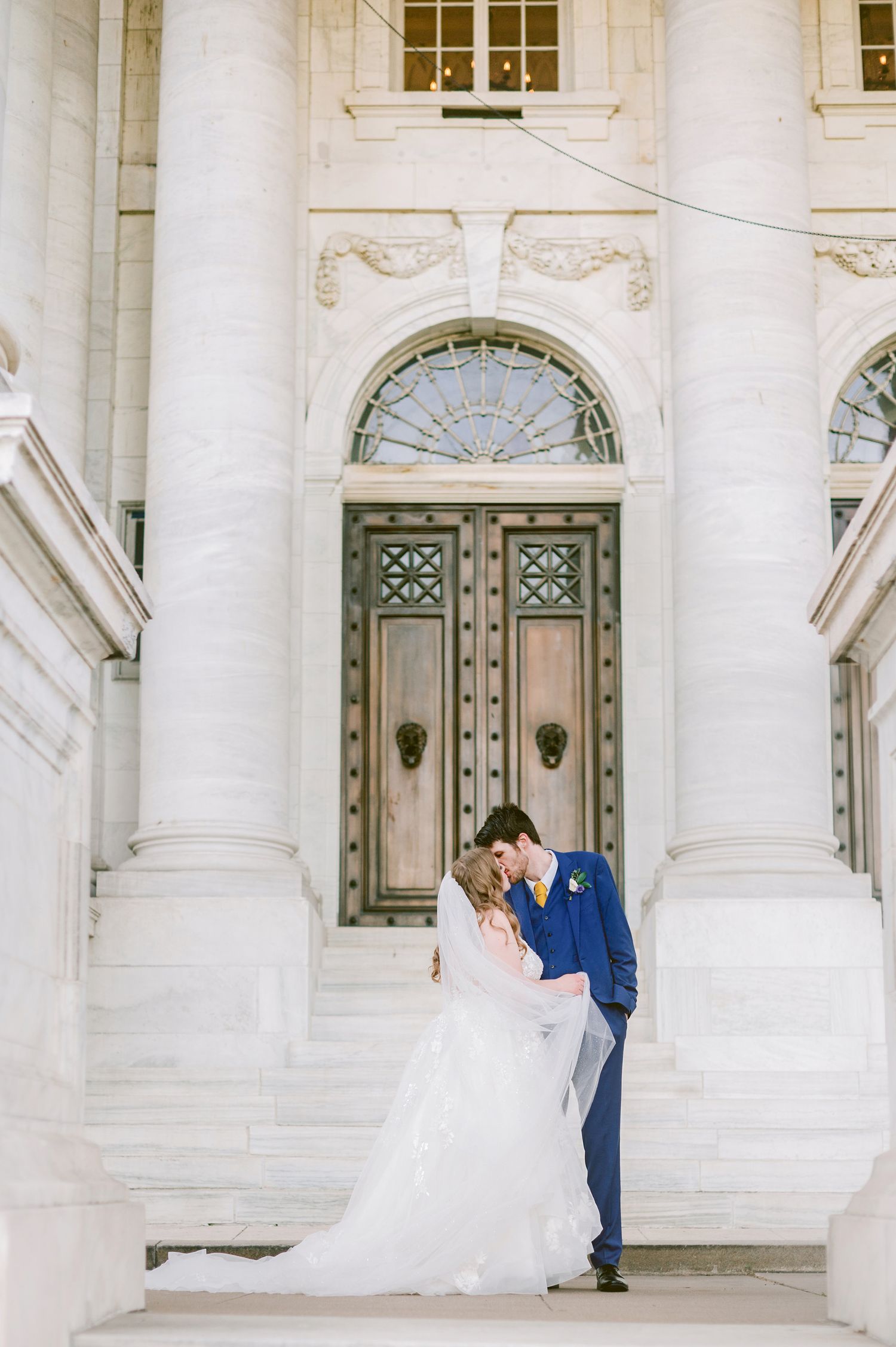Elegant wedding couple embraces on white marble steps in front of ornate architectural doorway with classical columns.