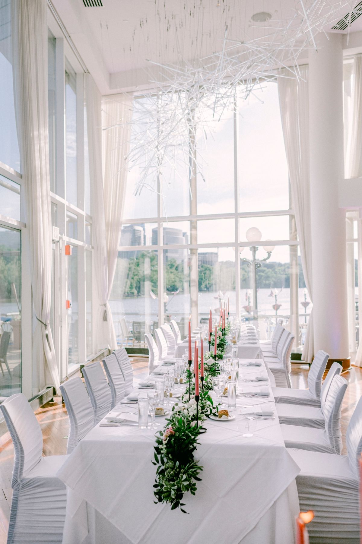 Elegant white reception venue with large windows, white chairs, and tables decorated with greenery centerpieces at Sequoia Restaurant in DC.