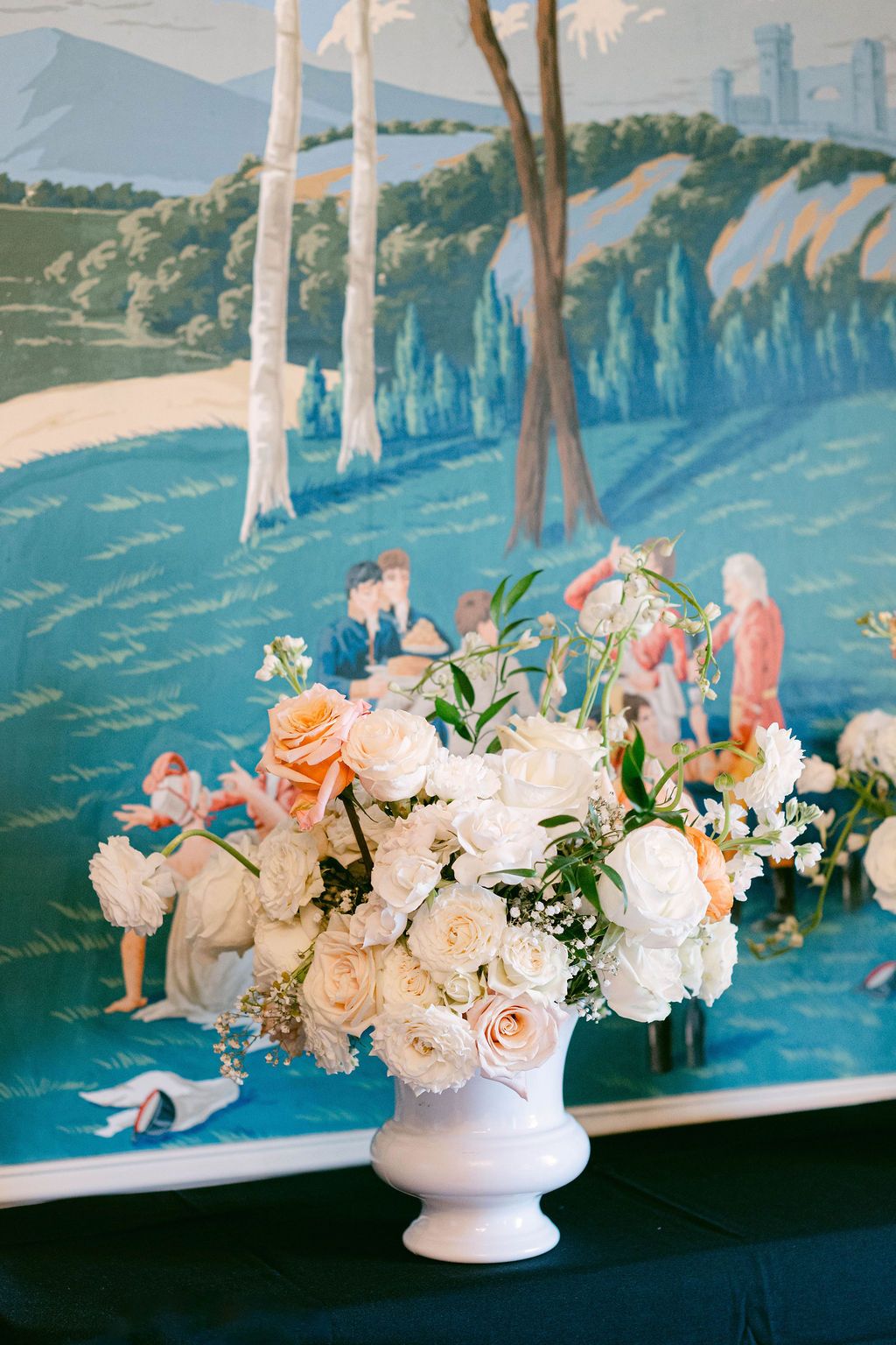 A series of elegant white and pink floral arrangements in white pedestal vases against a scenic painted wall backdrop.