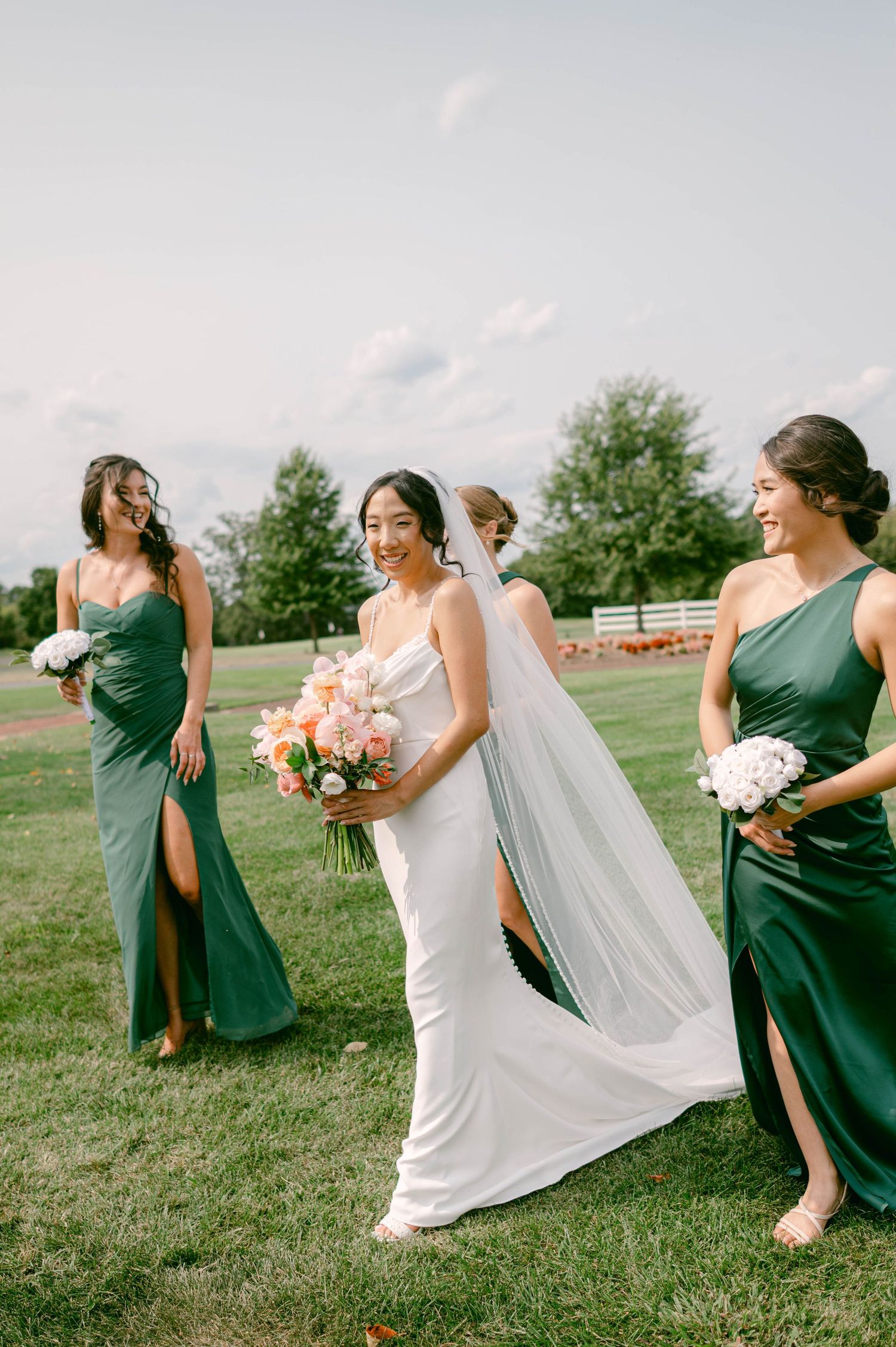 Wedding party in green bridesmaid dresses and white bridal gown pose playfully on grassy hill during outdoor event at Belmont Country Club.