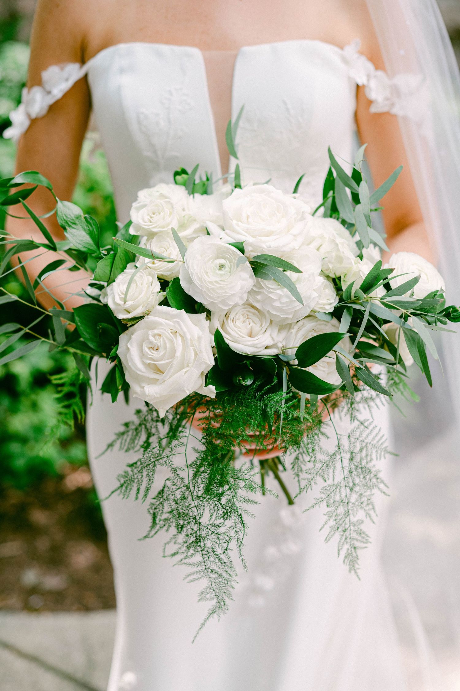 Elegant white rose and ranunculus bridal bouquet with trailing greenery held against a fitted wedding gown.