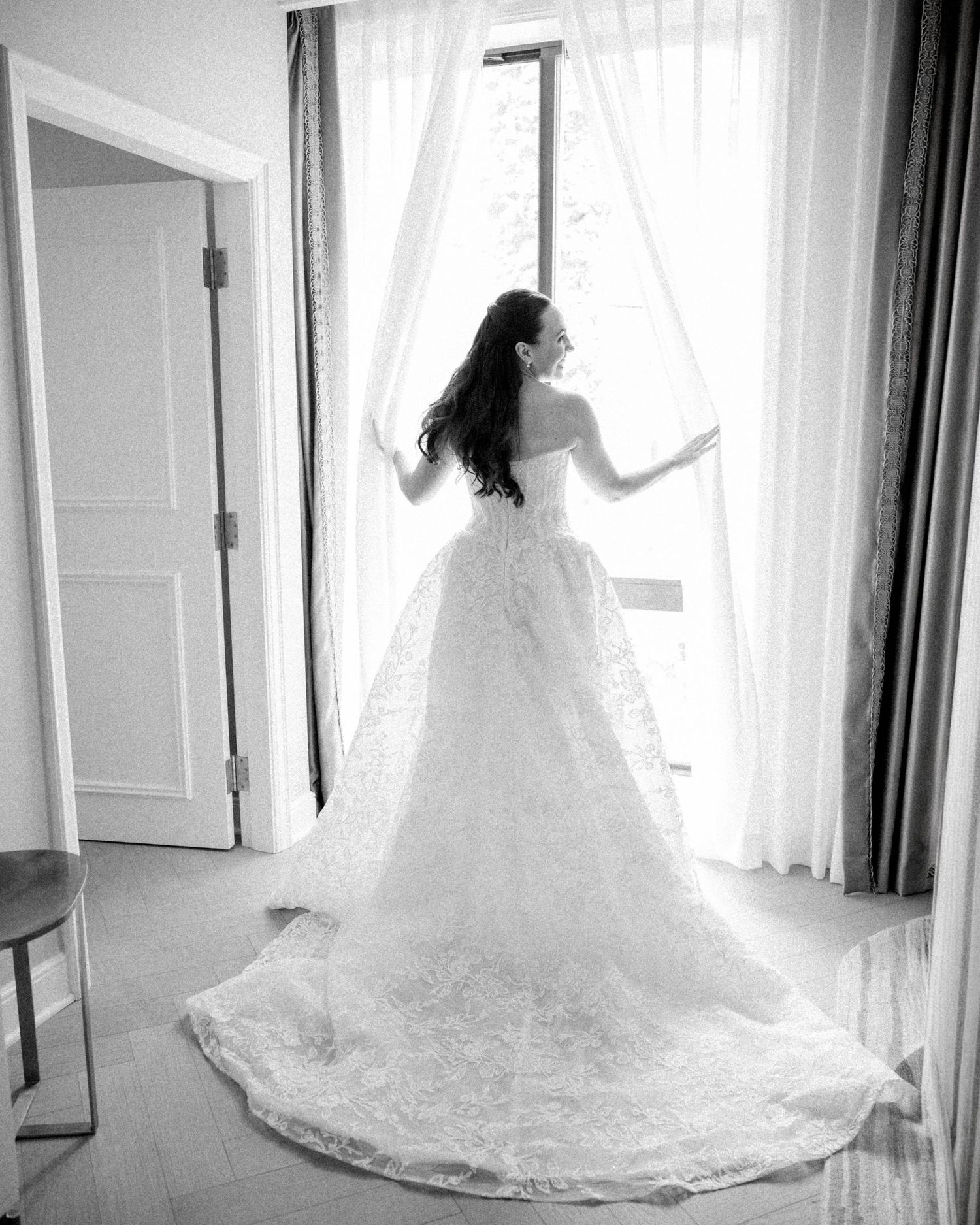 Black and white photo of a bride in a lace ball gown with long train standing at a window.