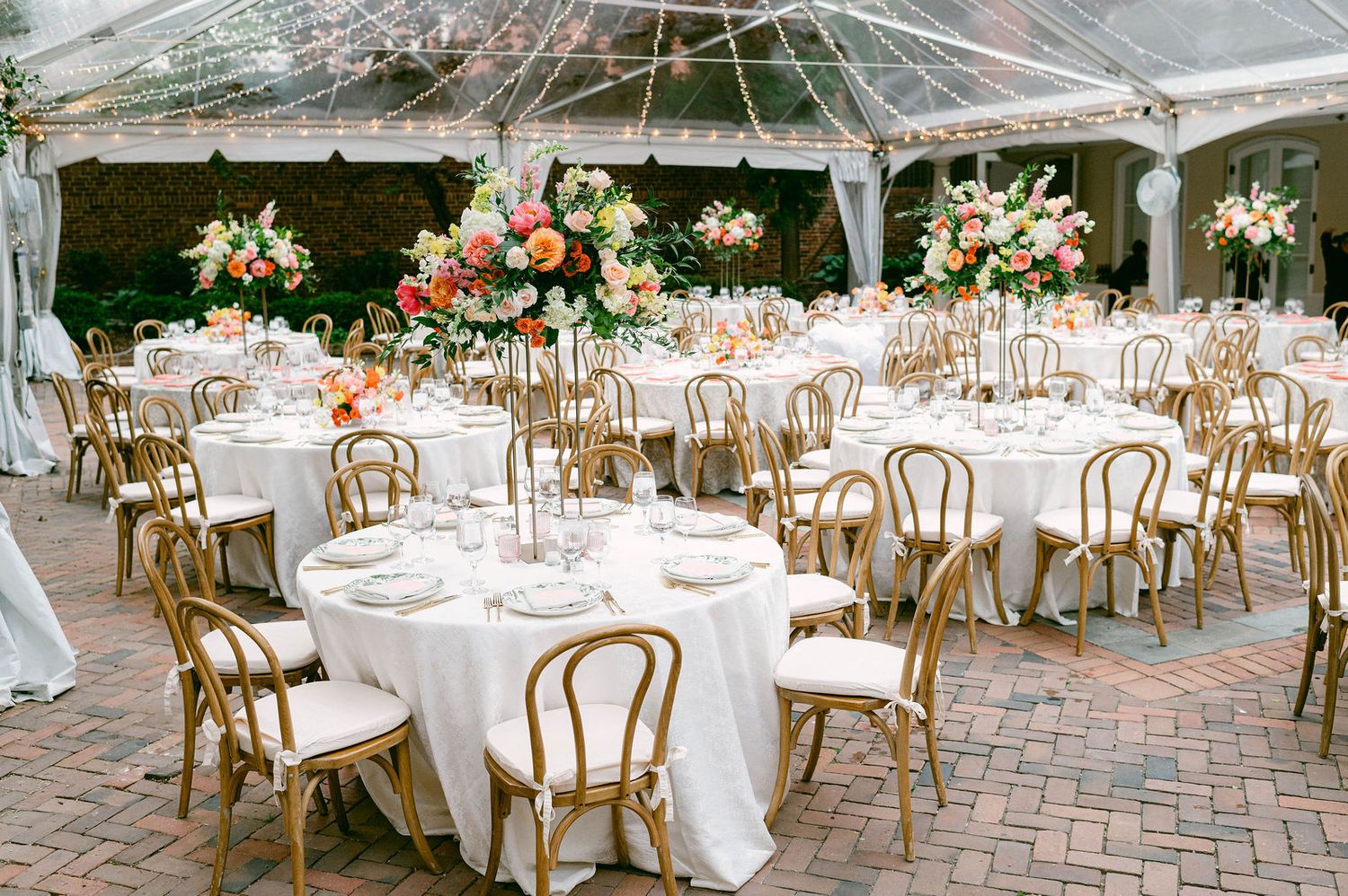 Elegant outdoor wedding reception setup with white tables, gold bentwood chairs and floral centerpieces under clear tent.
